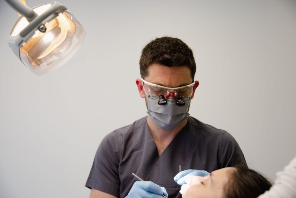 Dentist wearing a mask and magnifying loupes examines a patient under an overhead dental light